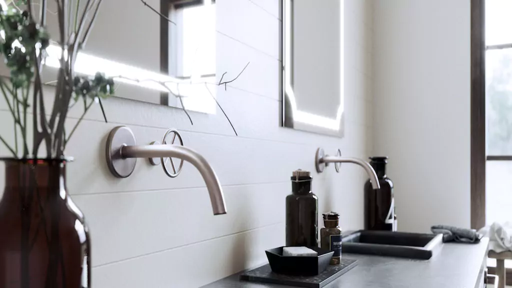 An illuminated mirror above a double sink with a brown bottle and flower vase in the bathroom.