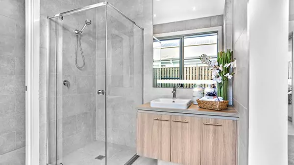 Glass-walled shower in a bathroom with a wooden cabinet sink and a lighted mirror above.